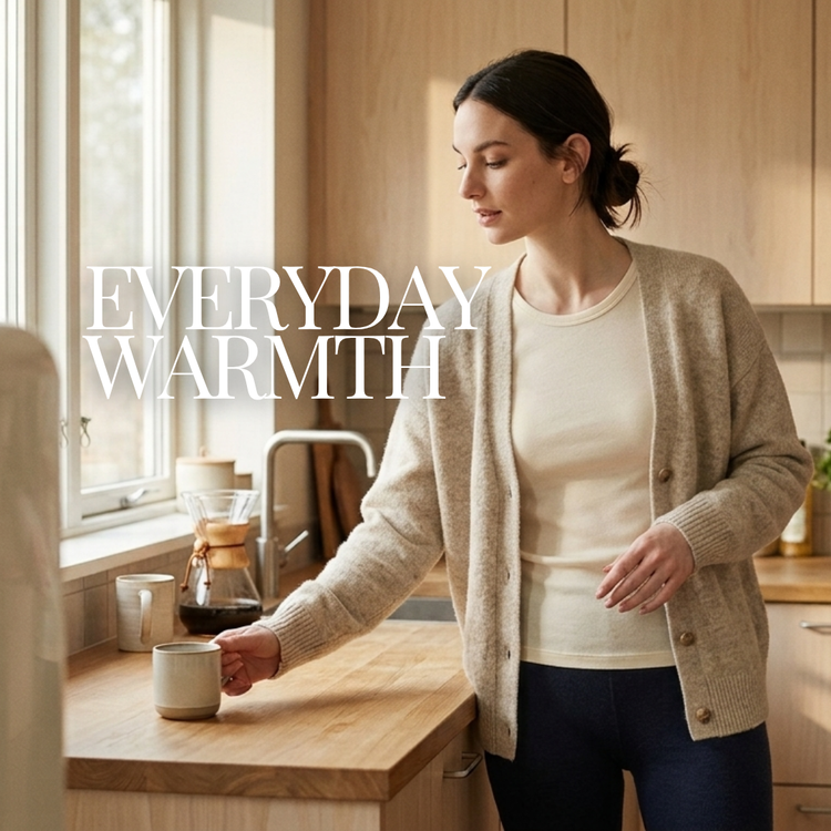 A woman stands in a warm, sunlit kitchen wearing a beige knit cardigan over a cream top and dark blue pants. She is reaching for a mug on a wooden counter, with large white text reading 'EVERYDAY WARMTH' overlaid on the left side of the image.