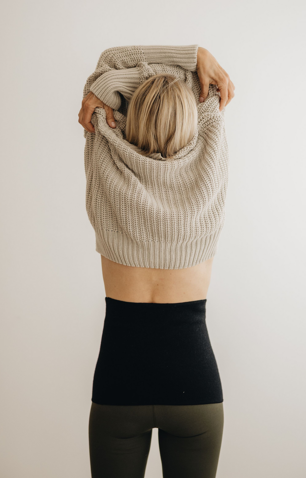 A woman viewed from behind wearing a chunky knit cowl-neck sweater in a light beige color, holding it up over her head. She is also wearing a black high-waisted back warmer and dark olive green leggings, set against a plain off-white background.